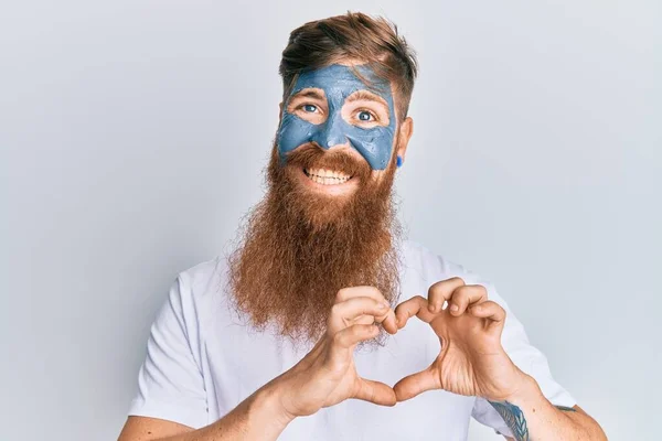 Young irish redhead man wearing facial mask smiling in love showing heart symbol and shape with hands. romantic concept.