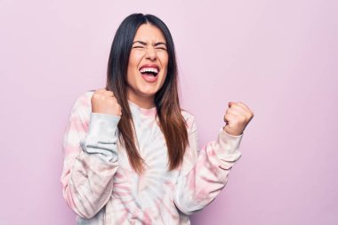 Genç, güzel esmer kadın, izole edilmiş pembe arka plan üzerine gündelik bir kazak giyiyor. Şaşırmış ve şaşırmış bir şekilde kolları havada ve gözleri kapalı bir şekilde başarıyı kutluyor.