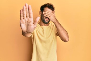 Sarı arka planda sarı tişört giyen, sakallı yakışıklı adam elleriyle gözlerini kapatıyor ve üzgün ve korku ifadeli hareketler yapmayı kesiyor. utangaç ve olumsuz kavram. 