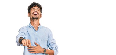 Gündelik kıyafetler giyen genç İspanyol adam sana gülüyor, eli vücudunun üzerinde kamerayı işaret ediyor, utanç ifadesi. 