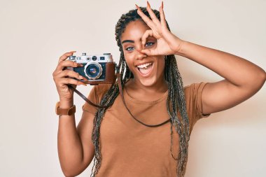 Saçları örülmüş, elinde antika fotoğraf makinesi olan genç Afro-Amerikalı kadın gülümsüyor ve gülümsüyor. 