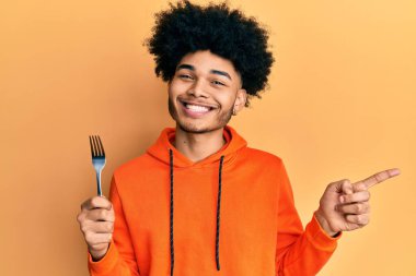 Afro saçlı, gümüş çatallı, gülümseyen, elini ve parmağını kenarı gösteren Afro Amerikalı genç bir adam. 