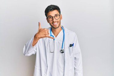 Young handsome man wearing doctor uniform and stethoscope smiling doing phone gesture with hand and fingers like talking on the telephone. communicating concepts. 