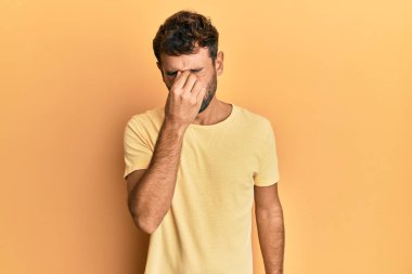 Sarı arka planda sarı tişört giyen sakallı yakışıklı adam yorgun burnu ve gözleri ovuşturup yorgun ve baş ağrısı çekiyor. Stres ve hüsran kavramı. 