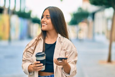 Genç İspanyol kız mutlu bir şekilde gülümsüyor. Akıllı telefon kullanıyor ve şehirde kahve içiyor..