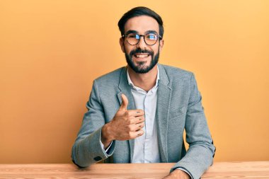 Young hispanic man working at the office doing happy thumbs up gesture with hand. approving expression looking at the camera showing success. 