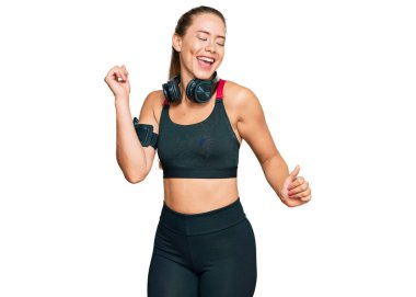 Güzel sarışın kadın spor kıyafetleri giyiyor ve kulaklık takıyor mutlu ve neşeli dans ediyor, gülümsüyor rahat ve kendinden emin bir şekilde müzik dinliyor. 