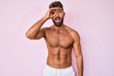 Latin asıllı genç adam, tişörtsüz duruyor. Şaşırmış yüz ifadesiyle şoka girmiş, parmaklarının arasından bakıyor. İnançsız ifade. 