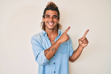 Yaz stili giyinmiş genç İspanyol adam gülümsüyor ve kameraya bakıyor iki eliyle ve parmaklarıyla kenarı işaret ediyor.. 