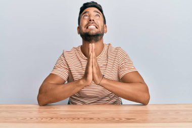 Yakışıklı İspanyol bir adam, gündelik giysiler içinde masada oturuyor, yalvarıyor ve dua ediyor. Yüzünde umut ifadesi var. Çok duygusal ve endişeli. Yalvarıyor. 