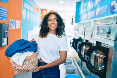 Kıvırcık saçlı, genç Afrikalı Amerikalı kadın çamaşırhanede iş yaparken mutlu gülümsüyor.
