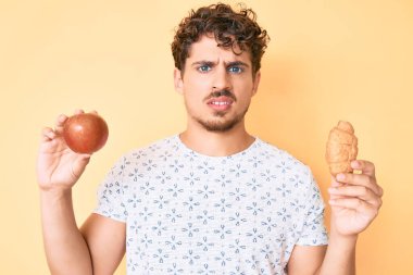 Kıvırcık saçlı, elma ve ayçöreği tutan, kafası karışık, genç, beyaz bir adam. şüphe kavramı. 