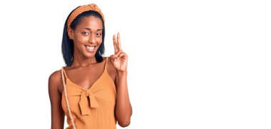 Genç Afro-Amerikalı kadın yaz kıyafeti giyip iki numarayı gösterip işaret ederken kendinden emin ve mutlu bir şekilde gülümsüyor.. 