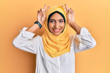 Genç esmer Arap kadın geleneksel başörtüsü takıyor komik ve çılgın poz veriyor parmakları tavşan kulağı gibi, neşeli gülümsüyor 