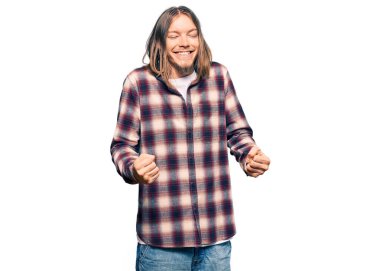 Uzun saçlı, hippi gömleği giyen yakışıklı beyaz adam kolları havada, gülümseyen ve başarı için bağıran hareketler yaparak çok mutlu ve heyecanlı. kutlama konsepti. 