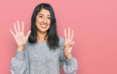Güzel Asyalı genç kadın gündelik giysiler giyer ve sekiz numaralı parmaklarıyla işaret ederken kendinden emin ve mutlu bir şekilde gülümser.. 