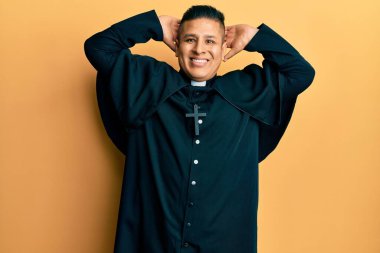 Genç Latin rahip sarı arka planda duruyor rahatlıyor ve esniyor, kolları ve elleri başının arkasında mutlu bir şekilde gülümsüyor. 