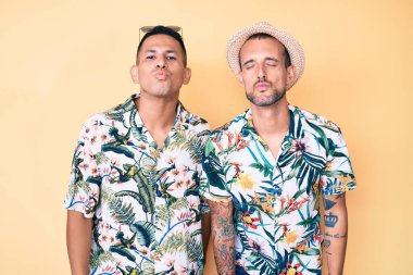 Genç eşcinsel çift, yaz şapkası ve Hawaii gömleği takmış kameraya bakıp havada öpüşerek güzel ve seksi oluyorlar. aşk ifadesi. 