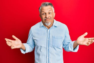 Orta yaşlı, gri saçlı, sıradan kıyafetler giyen, kafası karışmış, elleri ve kolları havada bir adam. şüphe kavramı. 