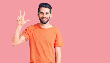 Sakallı, sıradan tişörtlü, üç numaralı parmaklarıyla gösteriş yapan, kendinden emin ve mutlu gülümseyen genç yakışıklı bir adam.. 