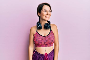 Kısa saçlı, spor giyimli, kulaklık takan, yüzünde gülümsemeyle yan yana duran esmer bir kadın. Kendinden emin gülen. 