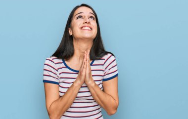 Latin asıllı genç bir kız, sıradan çizgili t-shirt giyip elleri bağlı yalvarıyor ve dua ediyor. Yüzünde umut ifadesi var. Çok duygusal ve endişeli. Yalvarıyor. 