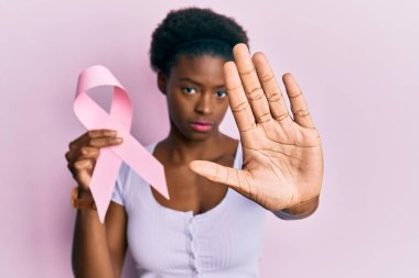 Genç Afro-Amerikalı kız elinde pembe kanser kurdelesiyle dur işareti yapıyor. Ciddi ve kendinden emin bir ifadeyle, savunma jesti. 
