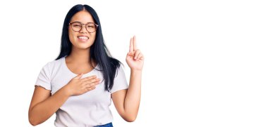 Genç, güzel Asyalı bir kız, günlük kıyafetler ve gözlük takıyor, gülümsüyor, elleri göğsünde ve parmakları havada küfür ediyor, sadakat yemini ediyor. 