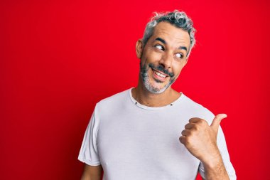 Orta yaşlı, gri saçlı, günlük beyaz tişört giyen, gülümseyen, gülümseyen ve baş parmağıyla yüzünü gösteren bir adam.. 