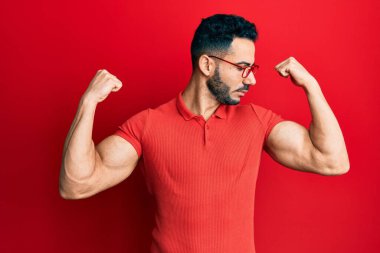 Sıradan kıyafetler ve gözlük takan genç İspanyol adam gururla gülümseyen kol kaslarını gösteriyor. fitness konsepti. 