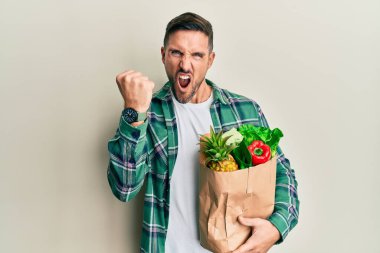 Sakallı yakışıklı bir adam elinde alışveriş torbalarıyla öfkeli ve kızgın bir şekilde yumruğunu kaldırıyor ve öfkeyle bağırıyor. öfke ve agresif konsept. 