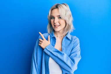 Gündelik kıyafetler giyen, neşeli gülümseyen, eli ve parmağı yana doğru olan genç sarışın kız. 