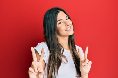 Genç esmer kadın kırmızı arka planda günlük beyaz tişört giyip kameraya bakıp zafer işareti yaparken gülümsüyor. İki numara.. 