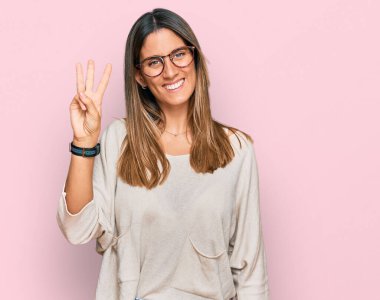 Gündelik kıyafetler giyen ve gözlük takan genç bir kadın üç numarayı gösterip işaret ederken kendinden emin ve mutlu bir şekilde gülümsüyor.. 
