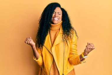 Orta yaşlı Afro-Amerikalı kadın yün kazak ve deri ceket giyiyor. Çok mutlu ve heyecanlı. Kolları havada, gülümsüyor ve başarı için çığlık atıyor. kutlama konsepti. 
