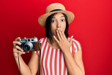 Yazlık şapkalı genç Latin kadın elinde antika kamerayla ağzını eliyle kapatıyor, şok geçiriyor ve hata yapmaktan korkuyor. şaşırmış ifade 