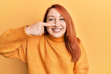 Sarı arka plan üzerine sıradan bir kazak giyen kızıl saçlı güzel bir kadın. El parmağı ve burunla işaret ediyor. Neşeli gülümsüyor. güzellik konsepti 