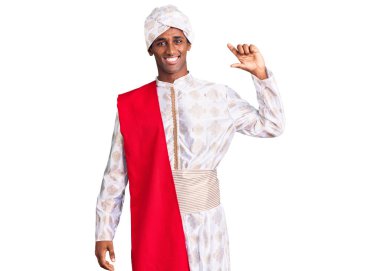 Geleneksel Sherwani sari giysileri giyen Afrikalı yakışıklı bir adam gülümseyen ve kendine güvenen el hareketleriyle küçük beden işareti yapan parmaklarıyla ve kamerasıyla. ölçüm kavramı. 