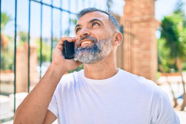 Orta yaşlı, kır saçlı, mutlu gülümseyen bir adam şehirde akıllı telefon kullanıyor.