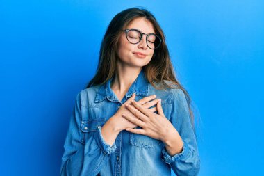 Gündelik kıyafetler ve gözlüklerle gülümseyen, elleri göğsünde, gözleri kapalı, yüzünde minnet ifadesi olan beyaz bir kadın. sağlık kavramı. 