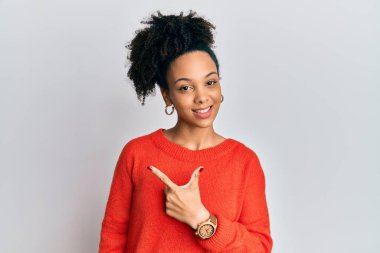 Genç Afro-Amerikan kızı, günlük giysiler giymiş, neşeli, yüzünde bir gülümsemeyle, mutlu ve doğal bir ifadeyle elini ve parmağını kenarı işaret ediyor. 