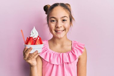 Güzel esmer küçük kız çilekli dondurma yiyor. Olumlu ve mutlu görünüyor. Kendine güvenen bir gülümsemeyle dişlerini gösteriyor. 