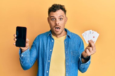 Poker kartı ve akıllı telefon tutan genç İspanyol adam boş ekranda korku ve şaşkınlık dolu bir ifade, korku ve heyecan dolu bir yüz sergiliyor.. 