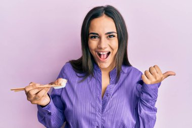 Genç esmer kadın nigiri suşisi yiyor yemek çubuklarıyla baş parmağını yana uzatıyor ve ağzı açık gülümsüyor. 