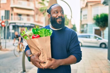 Sakallı Afrikalı Amerikalı bir adam elinde süpermarketten alınmış kağıt poşetler tutuyor.