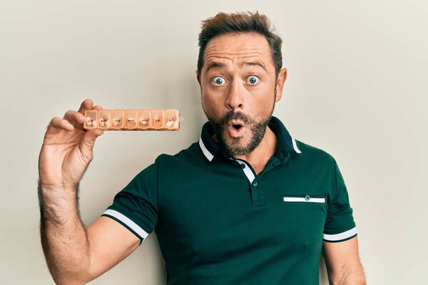 Middle age man holding pill organizer scared and amazed with open mouth for surprise, disbelief face 