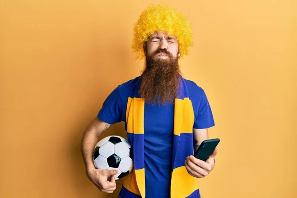 Redhead man with long beard football hooligan cheering game holding smartphone puffing cheeks with funny face. mouth inflated with air, catching air.
