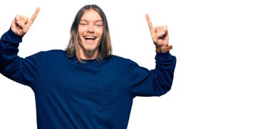 Uzun saçlı yakışıklı beyaz adam, sıradan bir süveter giyiyor. Gülümsüyor, şaşırıyor, parmaklarıyla işaret ediyor ve kollarını kaldırıyor.. 