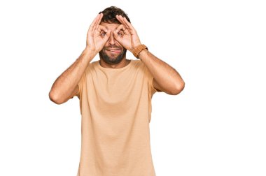 Sakallı, normal tişörtlü yakışıklı bir genç. Dürbün gibi jest yapıyor. Dilini çıkarıyor, gözleri parmaklarından bakıyor. çılgın ifade. 