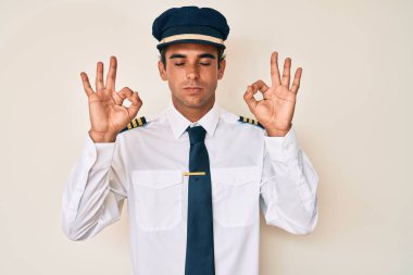 Uçak pilotu üniforması giyen, gözleri kapalı gülümseyen genç İspanyol adam parmaklarıyla meditasyon hareketi yapıyor. yoga konsepti. 